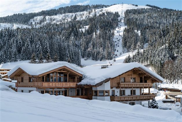 A charming mountain house in the snow with wooden cladding. In the background, snow-covered mountains and fir trees stretch out.