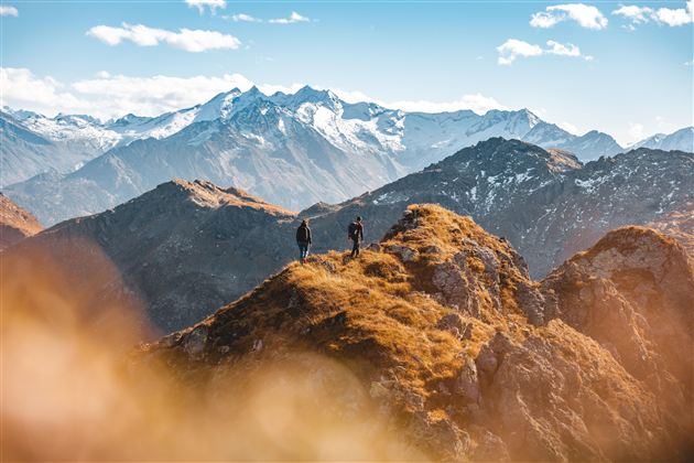Two hikers stand on a mountain peak with an impressive view of snow-covered mountains. The sky is blue with a few white clouds.