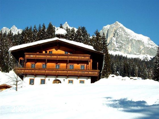 A cozy wooden house in the snow, surrounded by tall fir trees. Majestic mountains can be seen in the background.