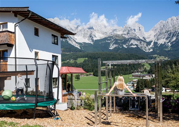 An idyllic house with a garden and playground, surrounded by majestic mountains.  
Children are happily playing on the swing and trampoline.