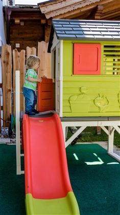 A small child is playing on a slide in front of a colorful playhouse. In the background, a wooden fence and green grass can be seen.