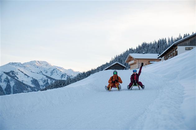 Two children are happily sitting in the snow and enjoying winter. In the background, snow-covered mountains and a mountain hut can be seen.