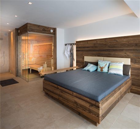 Ein modernes Schlafzimmer mit einem großen Bett und gemütlichen Kissen. Im Hintergrund befindet sich eine Sauna mit warmem Licht.