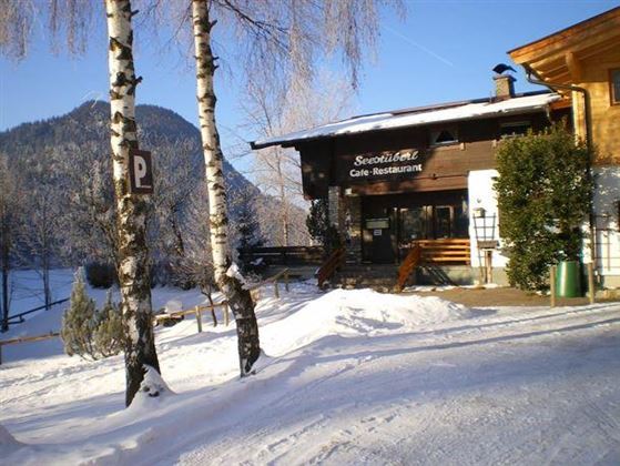 Ein schneeüberzogenes Restaurant mit einer rustikalen Holzfassade. Im Hintergrund sind verschneite Berge und strahlend blauer Himmel zu sehen.