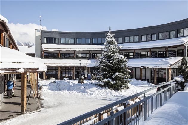 Ein ruhiger Innenhof mit schneebedeckten Gebäuden und einem großen Weihnachtsbaum. Der Himmel ist klar und die Umgebung wirkt einladend und winterlich.