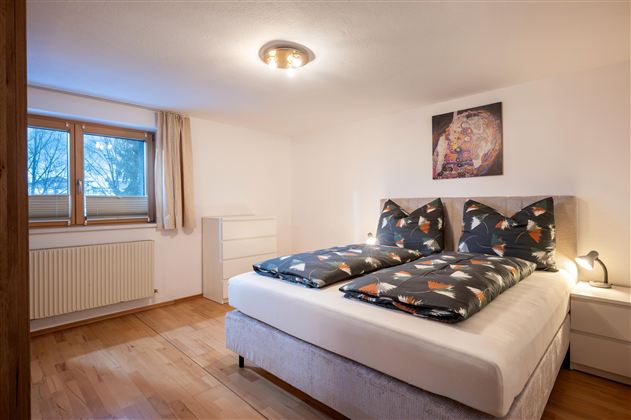 Ein gemütliches Schlafzimmer mit einem großen Bett und bunten Kissen. Im Hintergrund steht eine weiße Kommode und ein dekoratives Bild an der Wand.