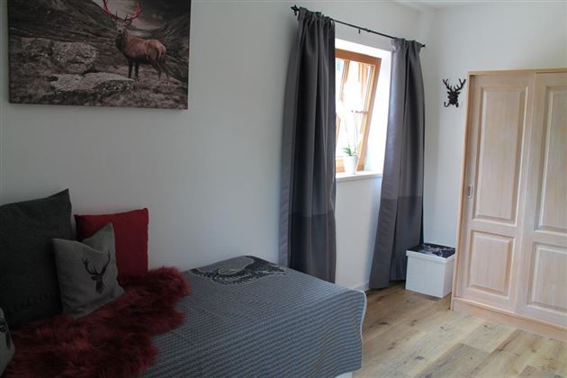 Ein gemütliches Schlafzimmer mit einem Bett und grauen Vorhängen. An der Wand hängt ein Bild eines Rehs, und der Boden ist aus Holz.