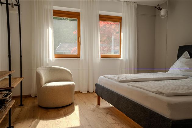 Ein helles Schlafzimmer mit zwei Fenstern und Tageslicht. Es gibt ein Bett und einen modernen Sessel.