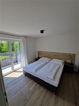 Ein helles Schlafzimmer mit einem Doppelbett und frischen Bettwäsche. Große Fenster bieten einen Blick auf die Natur draußen.