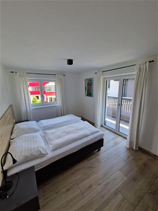 Ein helles Schlafzimmer mit einem Doppelbett und modernen Möbeln. Große Fenster bieten viel Tageslicht und Zugang zum Balkon.