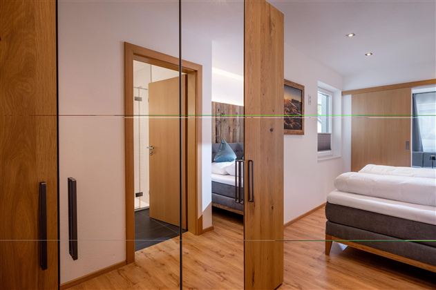 Ein modernes Schlafzimmer mit Holzmöbeln und einem großen Spiegel. Im Hintergrund ist ein bequemes Bett und eine Tür zu einem Badezimmer sichtbar.