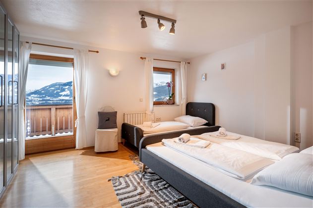 Ein gemütliches Zimmer mit mehreren Betten und hellem Holzfußboden. Die Wände sind mit Bildern dekoriert und es gibt große Fenster, die für ausreichend Licht sorgen.