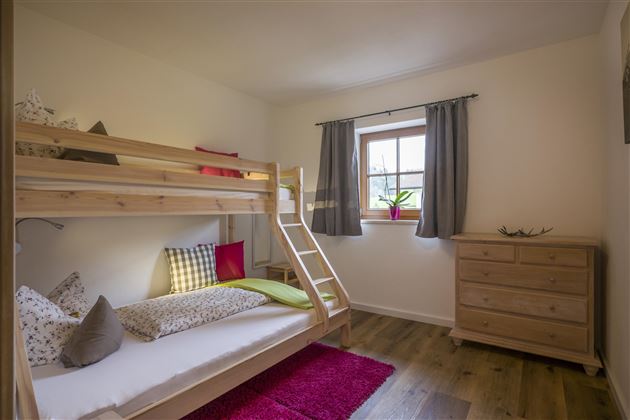 Ein gemütliches Kinderzimmer mit einem Etagenbett und einem Tagesbett. Das Zimmer hat ein Fenster mit einem Blick ins Freie und ist mit bunten Kissen dekoriert.
