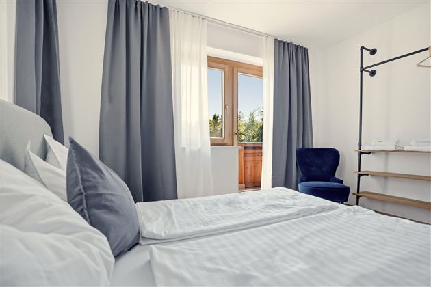Ein modernes Schlafzimmer mit einem Doppelbett und einem blauen Stuhl. Der Raum hat große Fenster mit Vorhängen und Blick nach draußen.