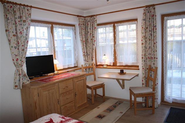 Ein gemütliches Zimmer mit Holzdekor, einem Fernseher und einem kleinen Tisch. Große Fenster lassen viel Licht herein und es gibt Stühle für Gäste.