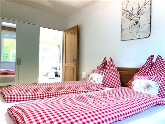 Ein gemütliches Schlafzimmer mit zwei Einzelbetten, die mit kariertem Bettzeug in Rot und Weiß ausgestattet sind. An der Wand hängt ein Bild eines Rehs, und der Raum wirkt hell und einladend.