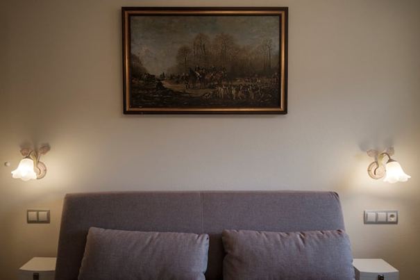 A cozy bedroom with a gray bed and a painting on the wall. Next to the bed, there are two wall lamps.