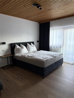 Ein gemütliches Schlafzimmer mit einem Doppelbett und zwei Kissen. Große Fenster lassen viel Licht herein und der Boden ist aus Holz.