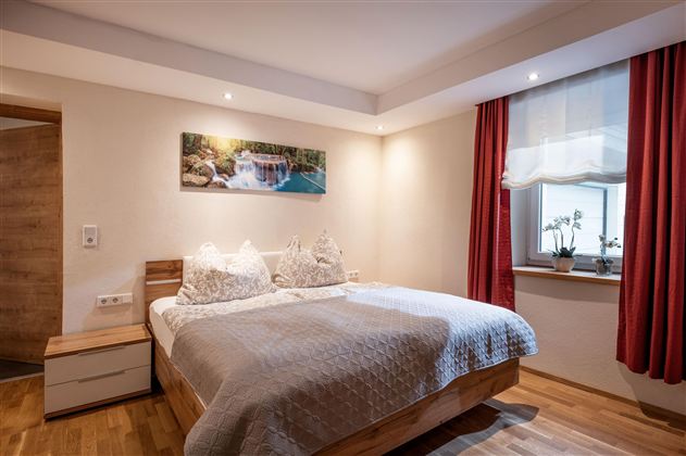 Ein modernes Schlafzimmer mit einem großen Bett und hellen Wänden. Ein Fenster mit Vorhängen und ein Bild an der Wand sorgen für eine gemütliche Atmosphäre.