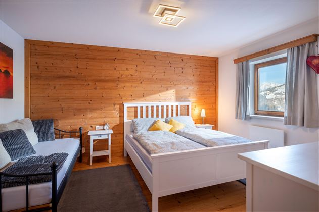 Ein gemütliches Schlafzimmer mit einem großen Bett und hellem Holzdekor. Ein Fenster lässt Tageslicht herein, und eine kleine Couch ergänzt den Raum.
