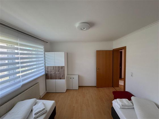 A simple and bright bedroom with two beds and wooden floor. There is a wardrobe and another room in the background.