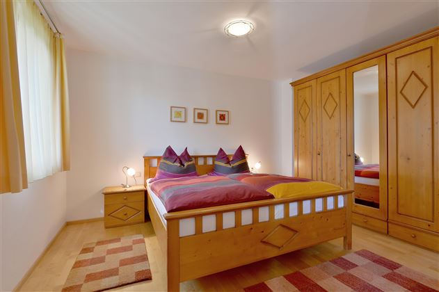 A cozy bedroom with a double bed and colorful cushions. The room is bright and inviting with a wooden wardrobe and a rug.