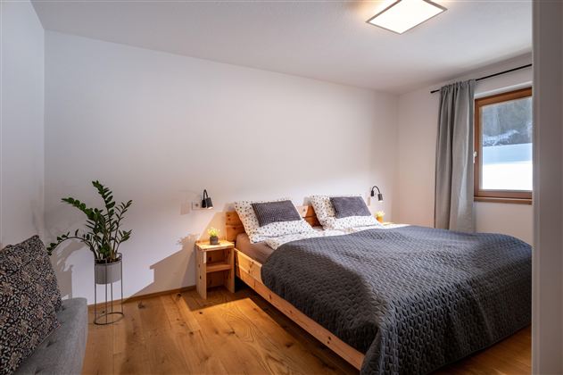 Een heldere slaapkamer met een groot tweepersoonsbed en witte muurverf. Naast het bed staan nachtkastjes met lampen en een raam zorgt voor daglicht.