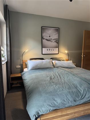 Een gezellige slaapkamer met een tweepersoonsbed en een zachte, blauwe dekbed. Aan de muur hangt een beeld met berglandschap en de tekst "SÖLL".