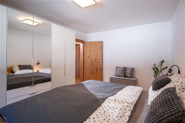 A modern bedroom with a large bed and a mirror wardrobe. The walls are light, and there are some plants for decoration.