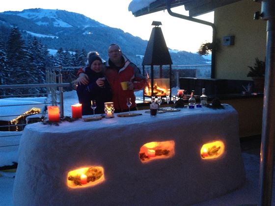 Ein gemütlicher Außenbereich mit einer Schneebar und stimmungsvollem Licht. Zwei Personen stehen zusammen und genießen die winterliche Atmosphäre in den Bergen.