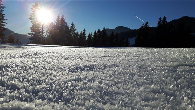 Eine verschneite Landschaft mit glitzerndem Schnee und einem strahlenden Sonnenuntergang. Im Hintergrund sind Berge und Bäume zu sehen.
