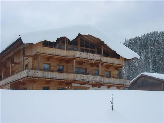 Ein zweistöckiges Holzhaus mit einem schneebedeckten Dach. Im Hintergrund sind verschneite Bäume zu sehen.