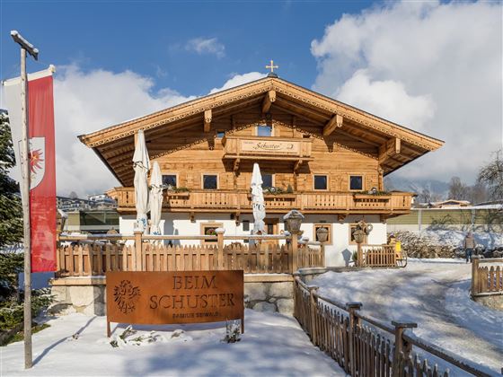 A traditional wooden house in Alpine style, surrounded by snow. In the foreground stands a sign reading "Beim Schuster".
