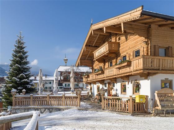 A cozy wooden house in the snow with balconies and a beautiful terrace. In the background, you can see additional buildings and a fir tree.