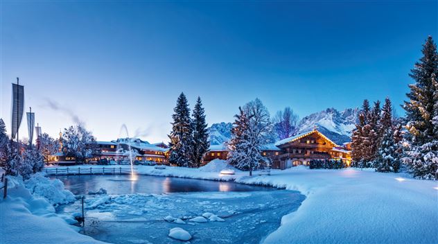 Eine malerische winterliche Landschaft mit schneebedeckten Bäumen und einem gefrorenen Teich. Im Hintergrund sind gemütliche Hütten und Berge unter einem klaren, blauen Himmel zu sehen.