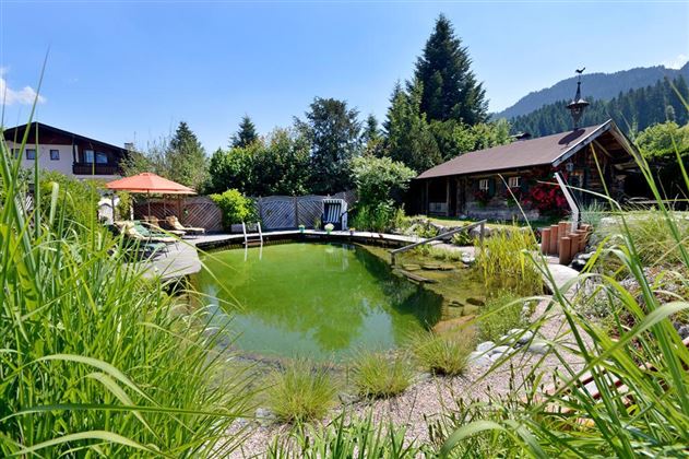 Ein idyllischer Naturpool umgeben von üppigem Grün und Blumen. Im Hintergrund ist ein gemütliches Holzhaus und ein Pavillon sichtbar.