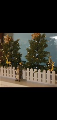 Zwei kleine, geschmückte Weihnachtsbäume stehen hinter einem weißen Zaun. Eine Figur steht vor dem Zaun und das Bild hat eine festliche Stimmung.