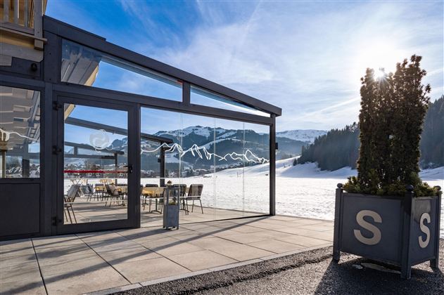 A modern building with large windows that offers a magnificent view of snow-capped mountains. On the terrace, there are tables and chairs, perfect for an outdoor stay.