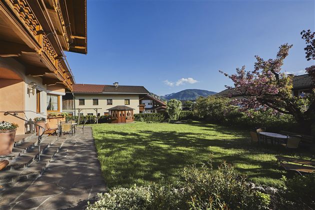 A well-maintained garden with green grass and blooming trees. In the background, picturesque mountains and charming houses can be seen.