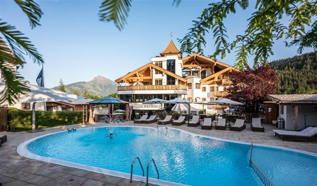 A pool in front of a charming hotel, surrounded by mountains and trees. The beautiful terrace invites you to relax.