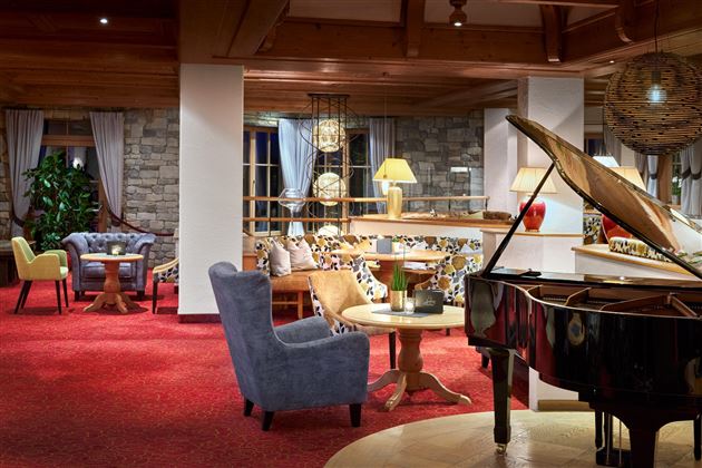 A stylish living room with a piano, cozy armchairs, and warm light. The walls are clad in stone and large windows let in plenty of daylight.