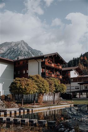 Ein malerisches Gebäude in den Bergen mit herbstlichen Bäumen und einem ruhigen Teich. Die Landschaft ist von einer beeindruckenden Bergkulisse umgeben.