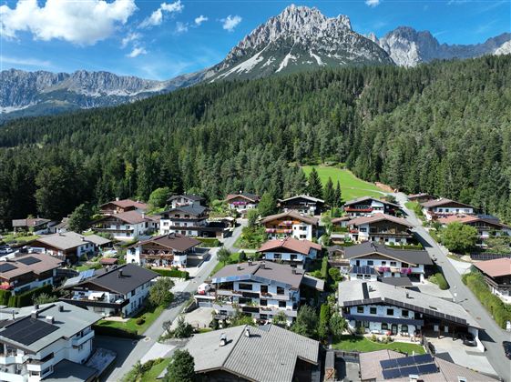 Eine malerische Berglandschaft mit vielen Holzhäusern und einer dichten Waldfläche. Im Hintergrund erheben sich majestätische Berg den Wilden Kaiser unter einem klaren blauen Himmel.