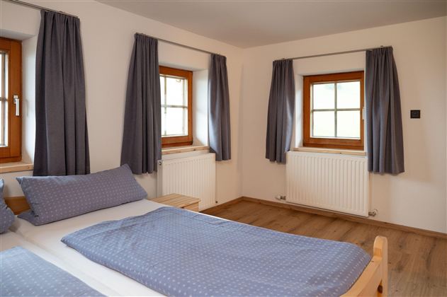 A bright bedroom with two windows and blue curtains. The bed is neatly covered with blue bed spreads.