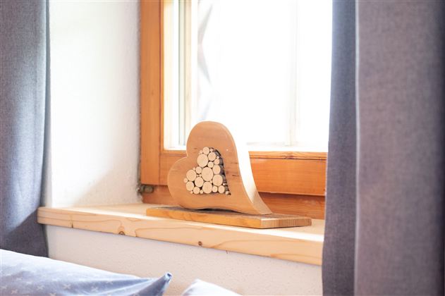 A wooden heart with a creative pattern of small sticks stands on a windowsill. In the background, curtains and a window can be seen.