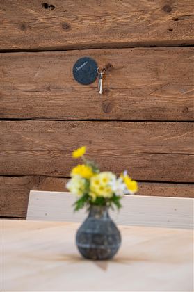 A small table with a vase full of flowers is in a rustic room. A key is hanging on a small sign on the wall.