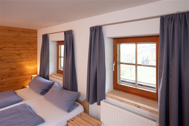 A cozy bedroom with two beds and blue cushions. Large windows with curtains let in plenty of light.