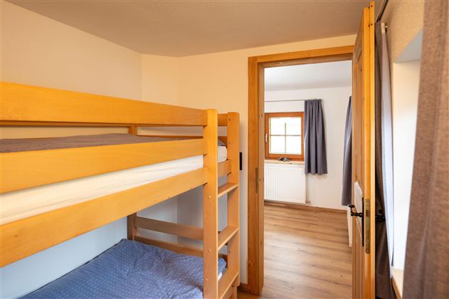 A simple room with a wooden bunk bed and a window. There is a bright wooden floor and a cozy entrance to another room.