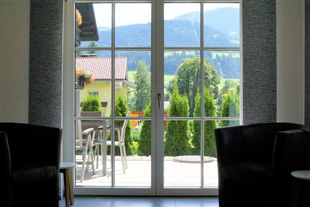 Een uitzicht door een raam op een groene landschap met bergen op de achtergrond. Voor het raam staan twee donkere stoelen.