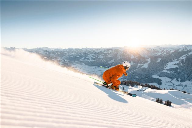 A skier in orange clothing glides down a snow-covered slope. In the background, impressive mountains stretch under a clear sky.